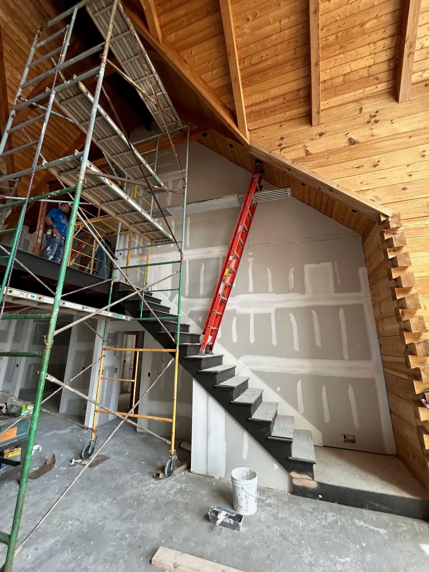 Drywall finishing on staircase wall with exposed wood ceiling, scaffolding